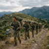 Soldiers in camouflage uniforms carrying rifles and backpacks hiking on a rocky mountain trail MTP Bergen